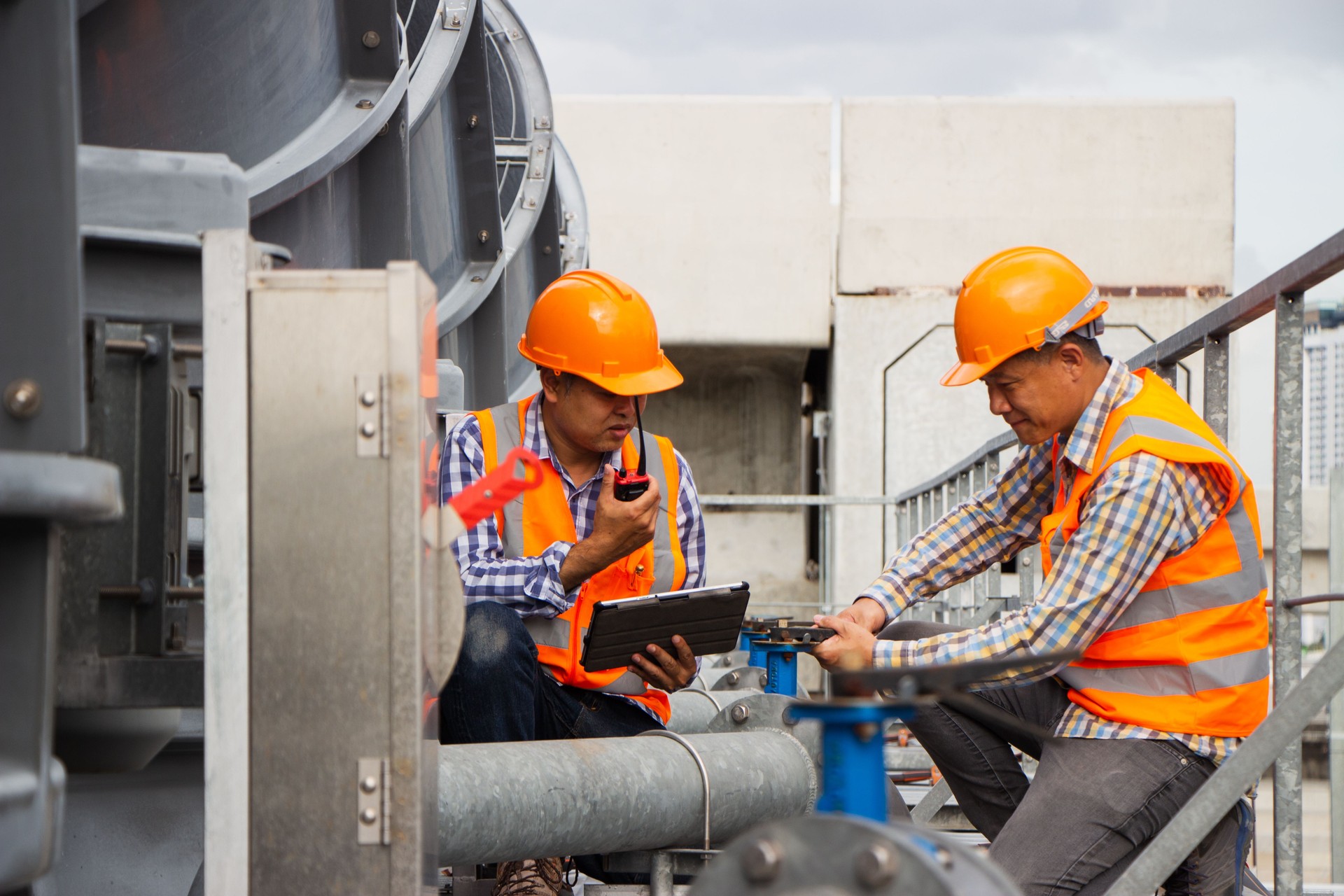 Asian male engineer inspecting pipe fittings and chillers, refrigeration plant refrigeration plant in  factory. male mechanic wearing helmet and Reflective Safety Vest uses laptop, tablet, equipment with sets of cooling towers in conditioning systems back